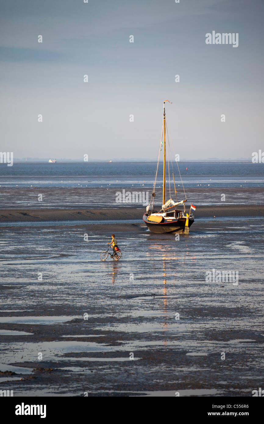 The Netherlands, Nes, Ameland Island, belonging to Wadden Sea Islands. Sailing boat on mud flat in harbour. Woman and bicycle. Stock Photo