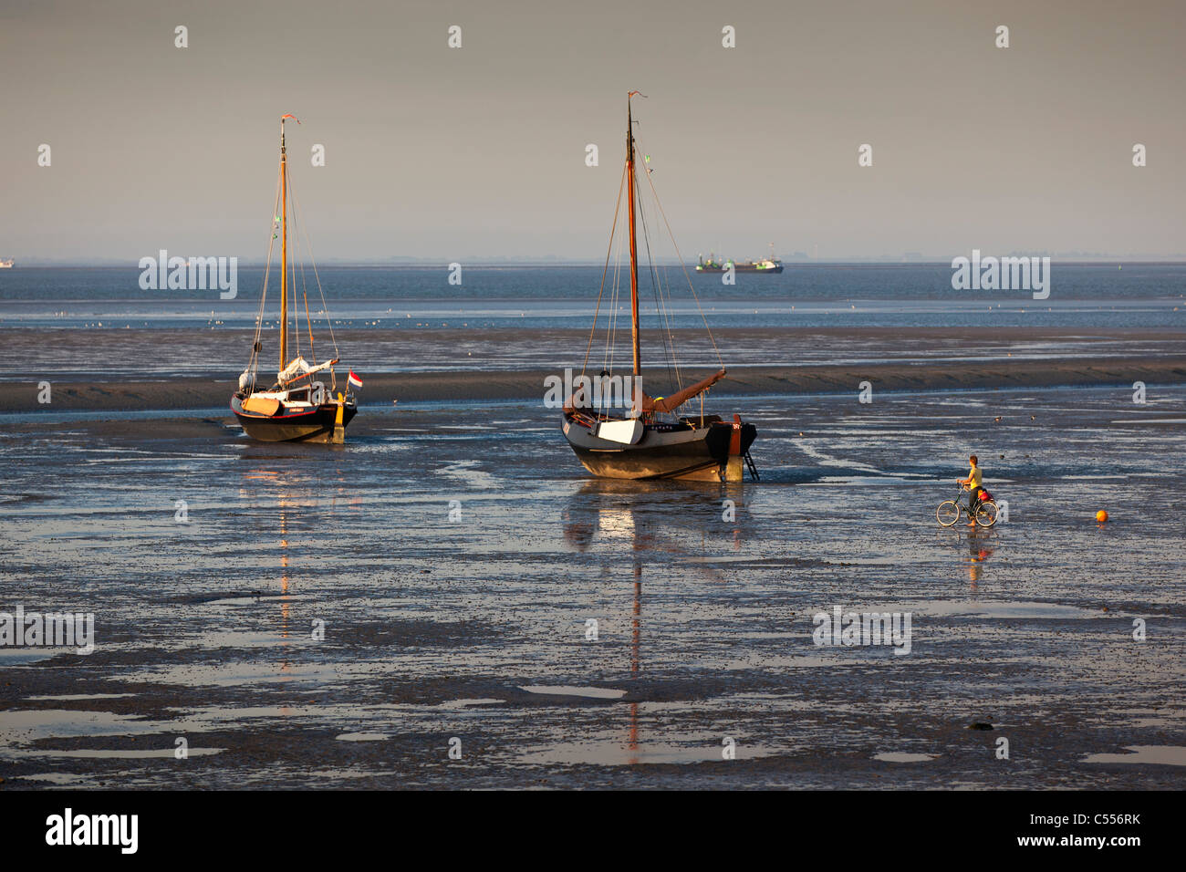The Netherlands, Nes on Ameland, Island belonging to Wadden Sea Islands. Woman with bicycle walking on mud flat. Sailing boats. Stock Photo