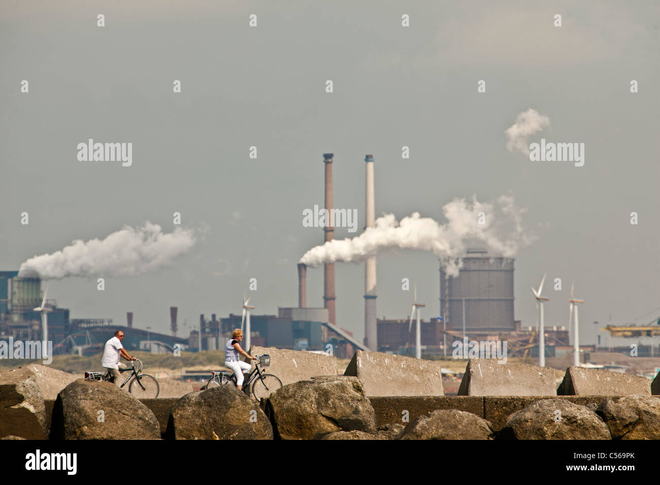The Netherlands, IJmuiden, Woman and man cycling on pier. Background Tata Steel factory, blast furnaces. Stock Photo