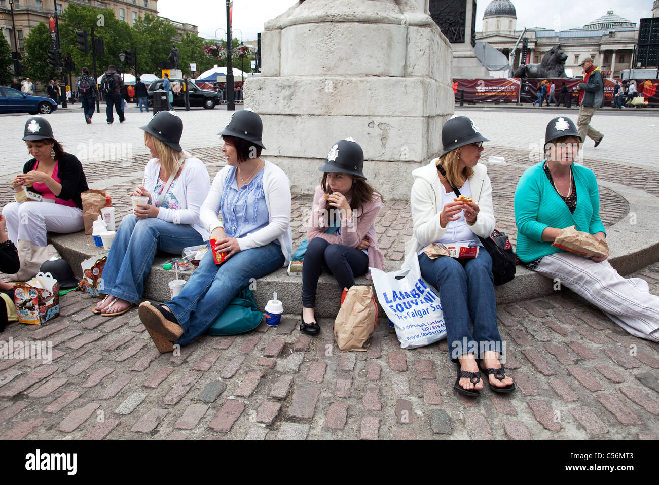 Group tourists eat their McDonalds in Trafalgar Square, London. All wearing tourist gift plastic policeman's helmets. UK. Stock Photo