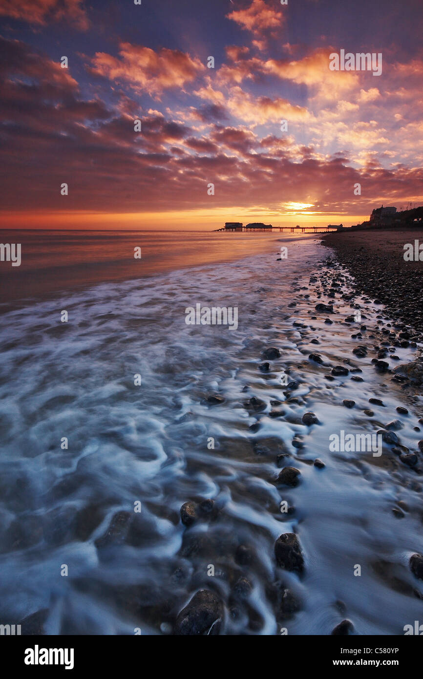 Looking along the shoreline at the sunrise behind Cromer pier on the Norfolk coast Stock Photo
