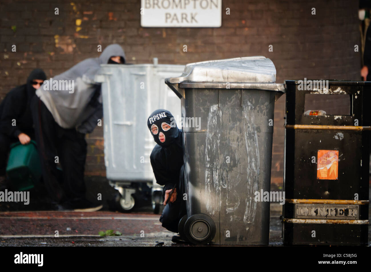Rioter wearing balaclava with initials 'FTPSNI' hides behind a dustbin with a rock in his hand to throw at police. Two rioters behind carry a crate of petrol bombs to throw Stock Photo