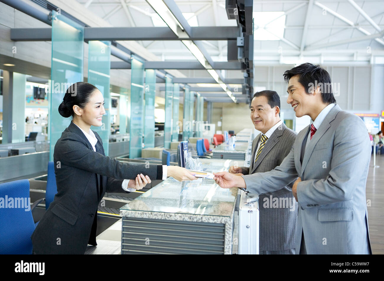 Businesswoman giving passport and airline ticket to young businessman while mature businessman looking at check-in counter Stock Photo