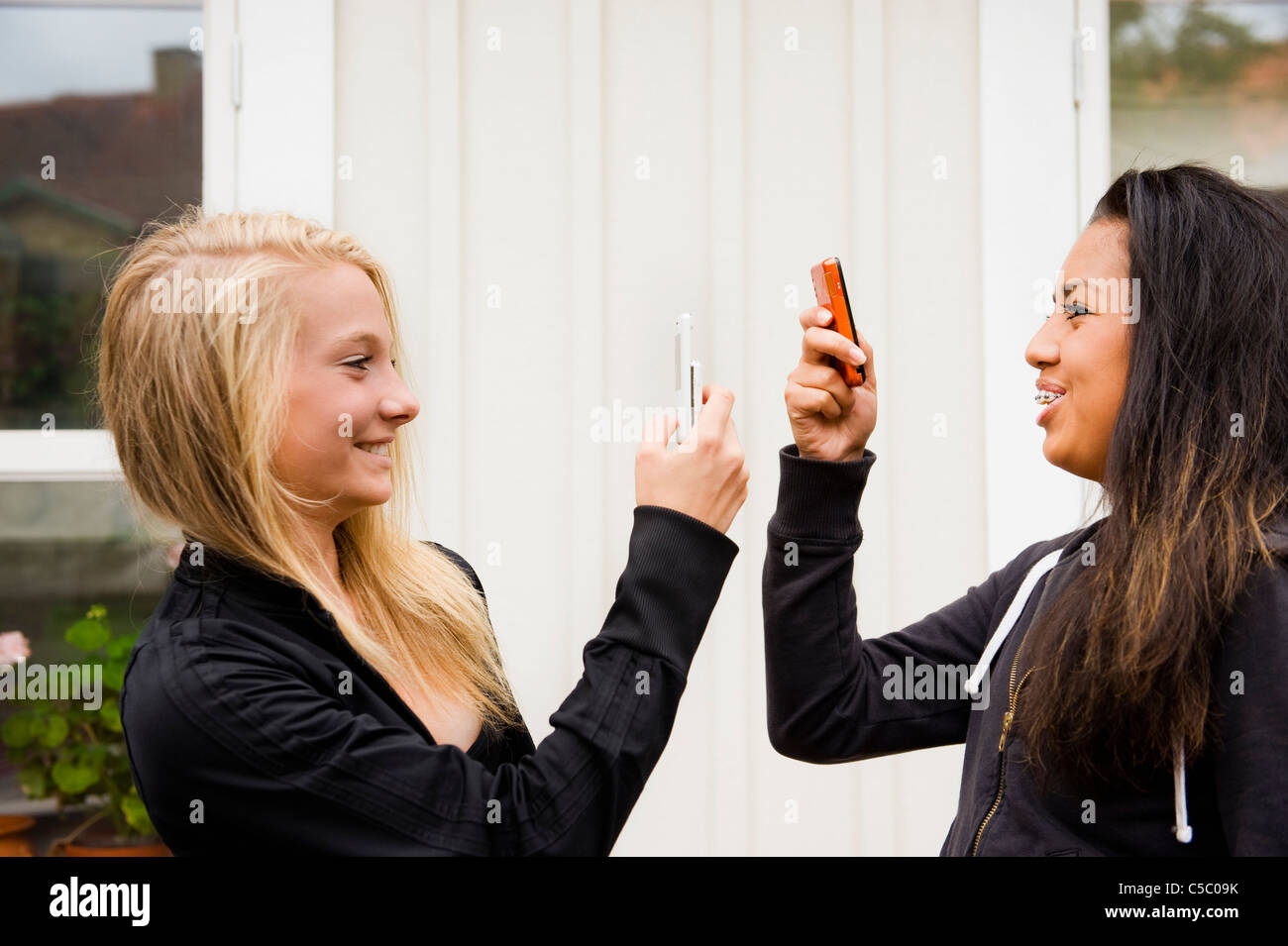 Side view of two multiracial teenage girls photographing each other with mobile phones Stock Photo