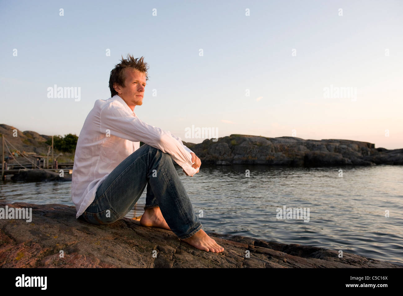 Side view of a thoughtful young man sitting on cliff by the sea against clear sky Stock Photo