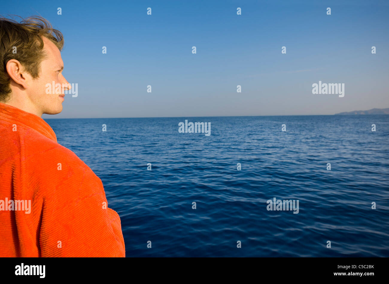Young man in orange bathrobe looking out at the peaceful sea against clear sky Stock Photo