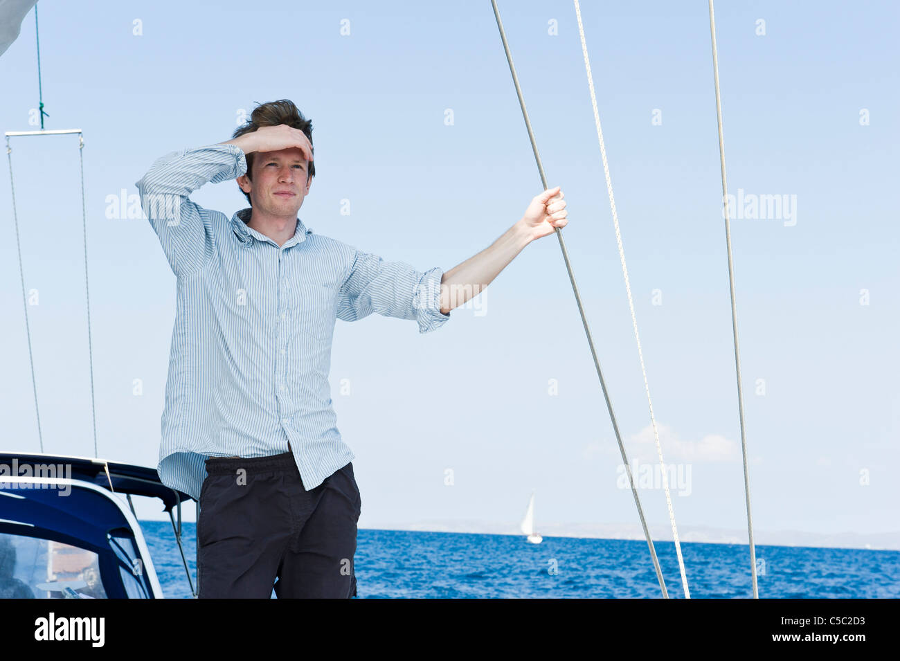 Young man shielding eyes on yacht against blue sea and clear sky Stock Photo