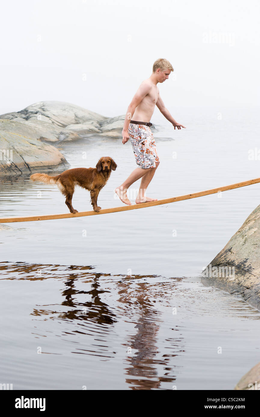 Side view of a shirtless boy and dog walking on a plank over the water Stock Photo