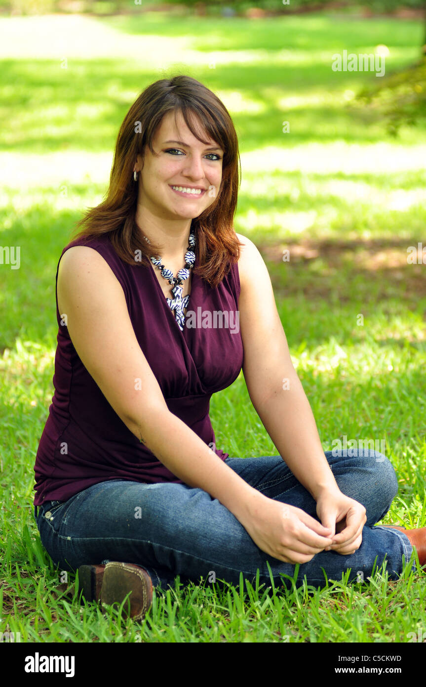 A pretty Hispanic woman is sitting on the grass looking into the camera. Stock Photo