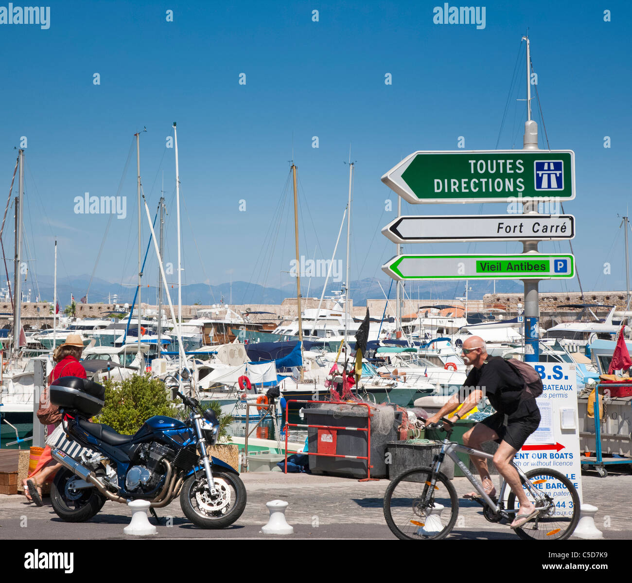 Cyclist and motorbike with direction sign and boats behind, Antibes, france Stock Photo