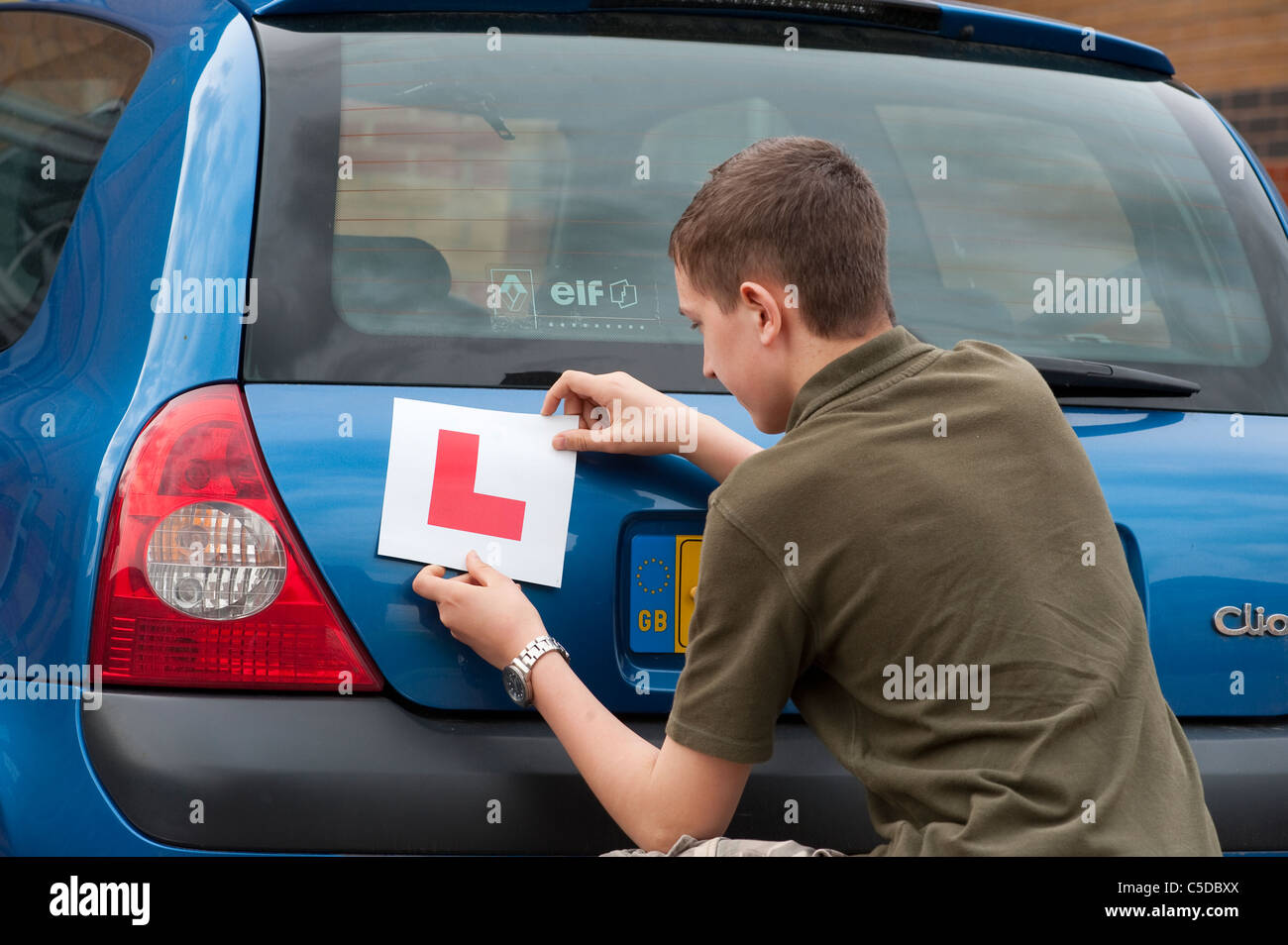 Teenage boy fixing an L plate to the back of a Renault Clio before a driving lesson. Stock Photo