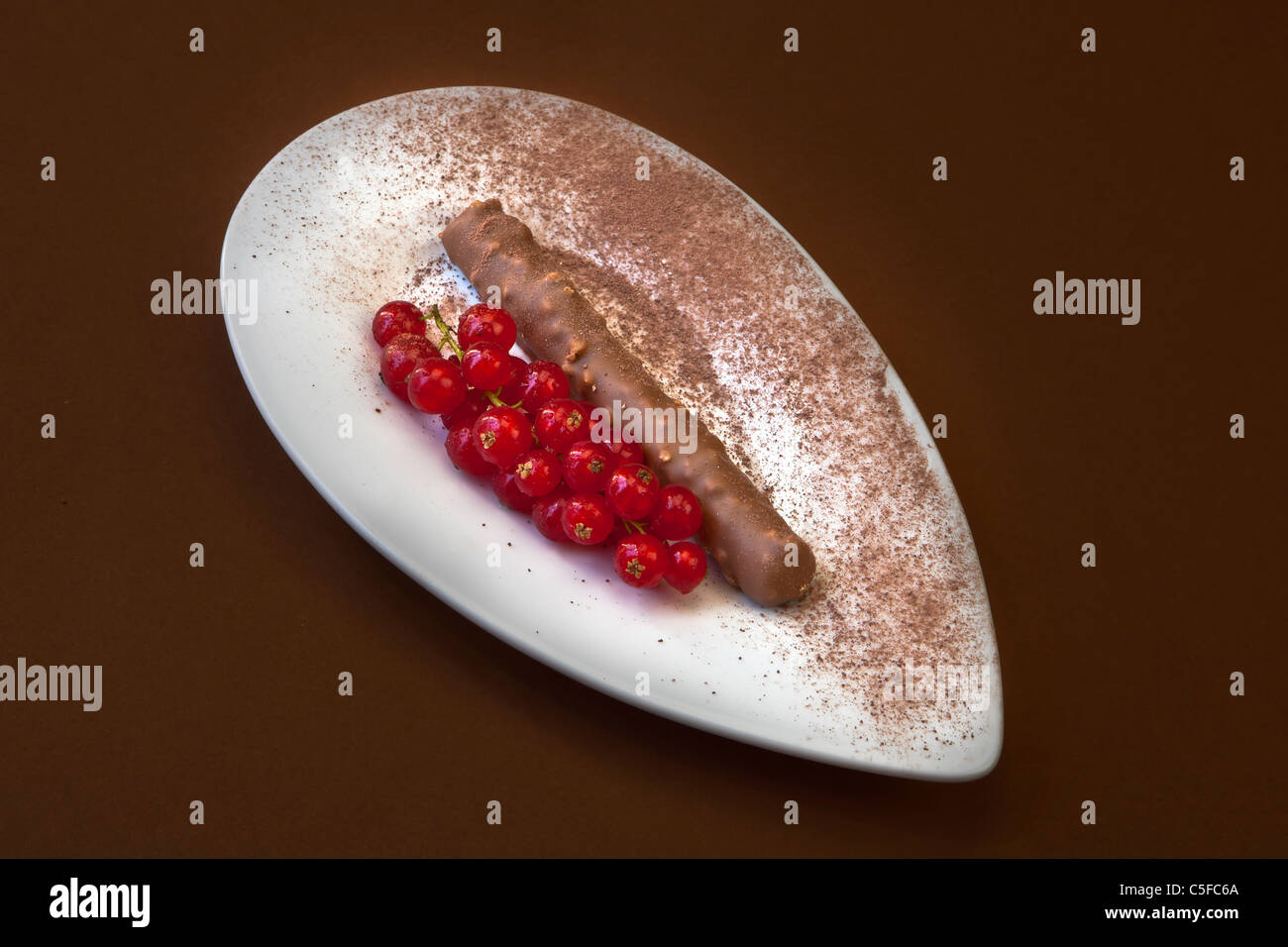 a long chocolate praline on a white plate with cocoa powder and red currants Stock Photo