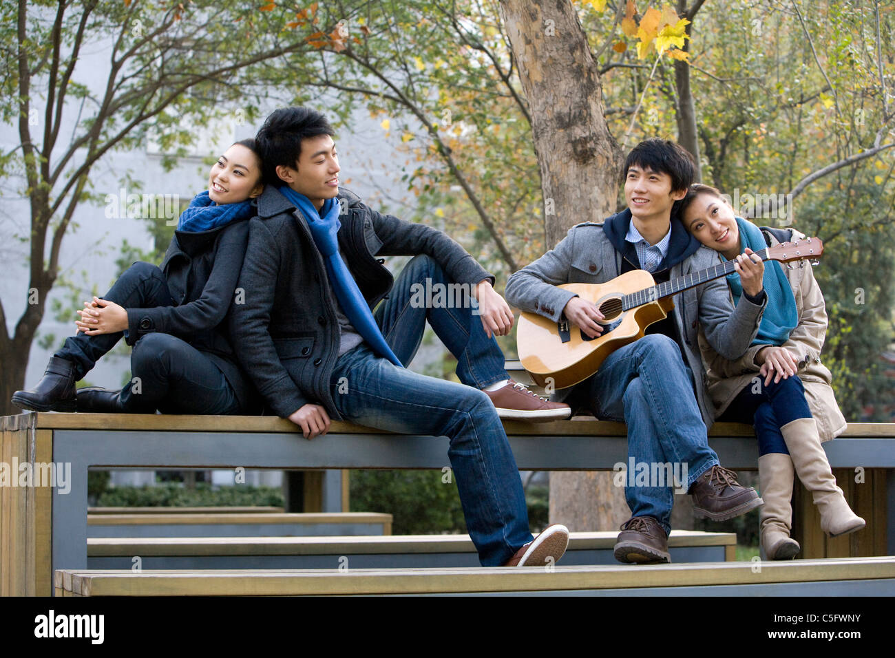 Two couples sit on a park bench and listen to a friend play guitar