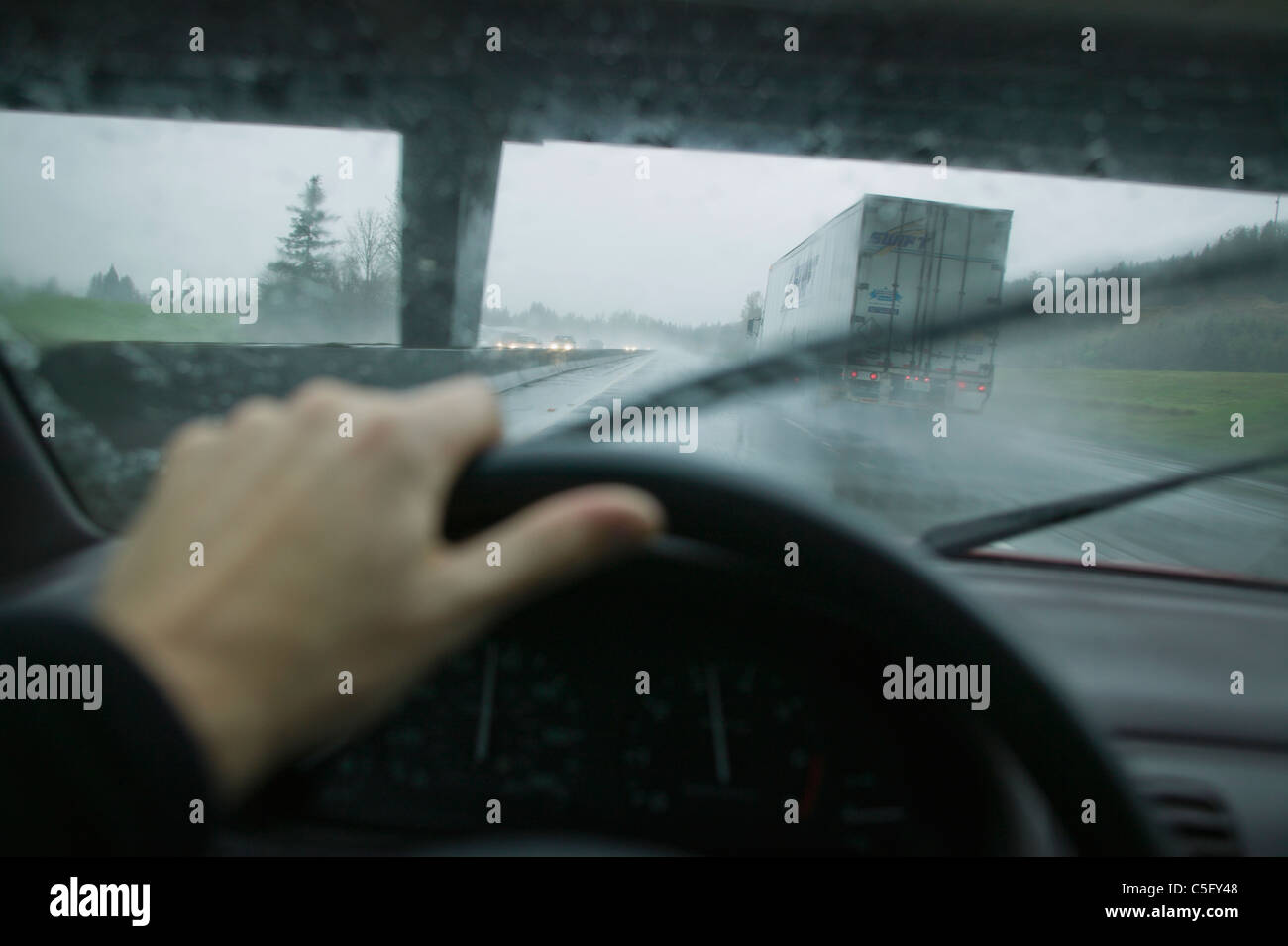 A first person perspective of driving in an automobile next to a truck trailer on a rainy day while passing below a overpass. Stock Photo