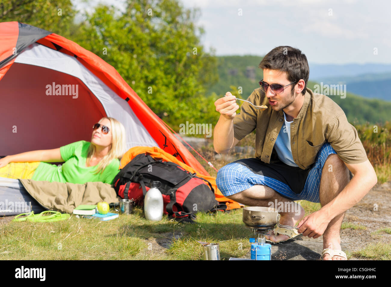 Happy camping couple with tent backpack cooking in sunny countryside Stock Photo