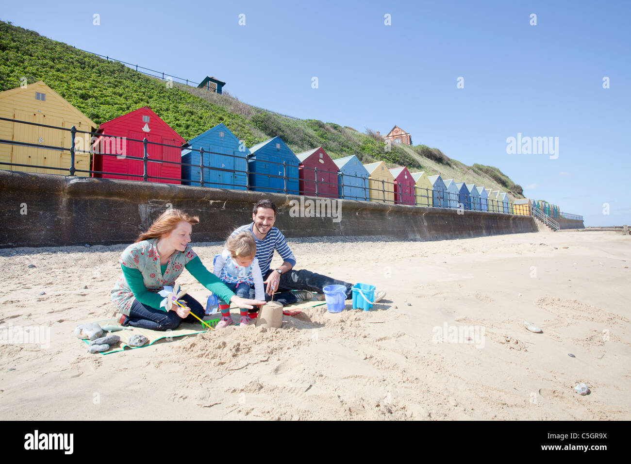 Young family building sand castles on the beach in front of beach huts at Mundesley on the Norfolk Coast Stock Photo