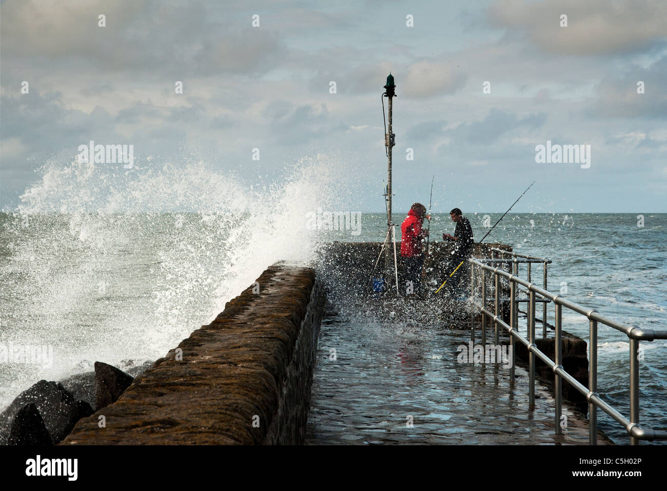 Big wave crashes over the sea defenses and splashes two sea fishermen at Lynton on the North Devon Coast, England, UK Stock Photo