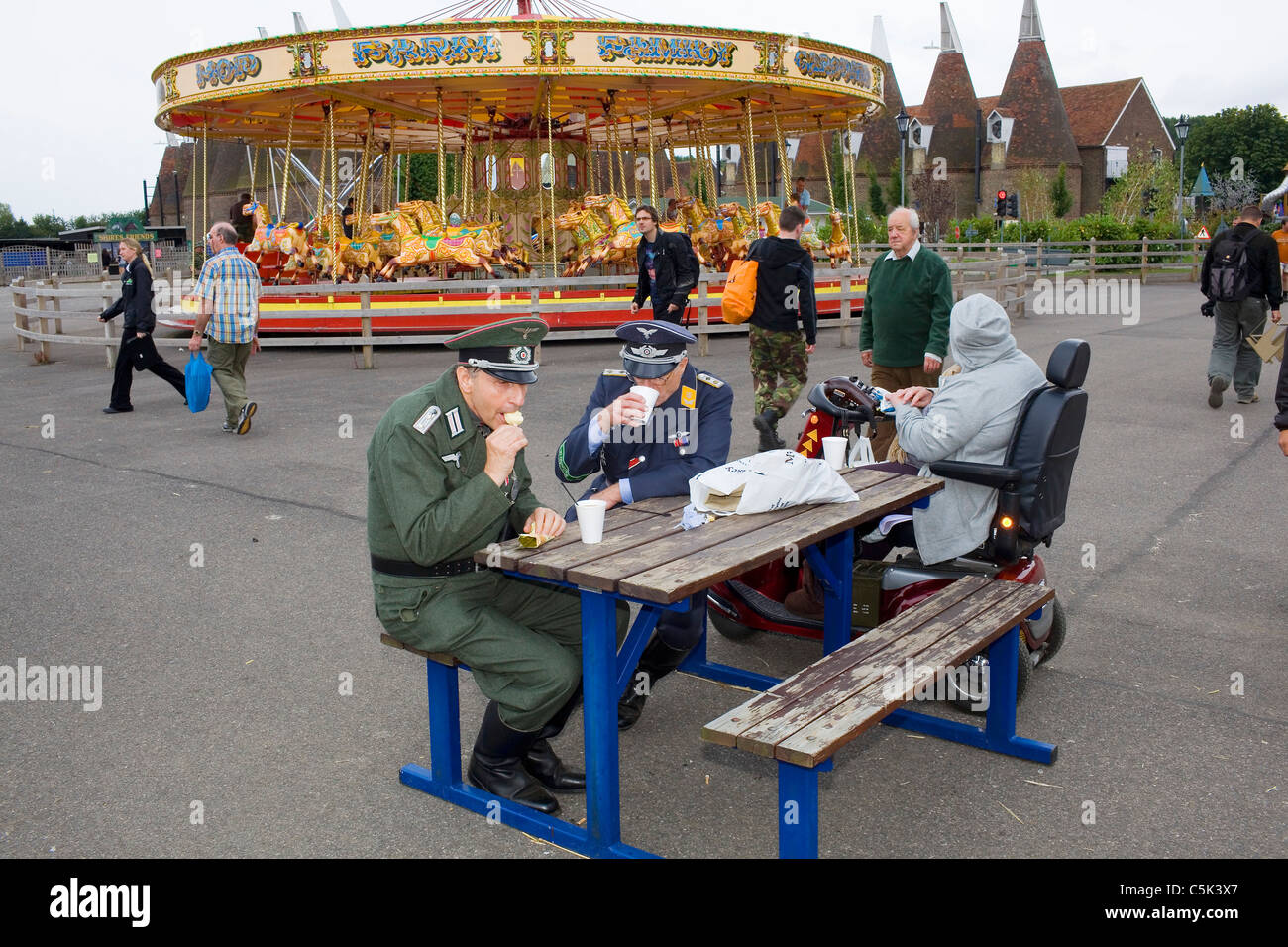 Two men wearing the uniform of German soldiers eat an ice cream by a carousel. Stock Photo