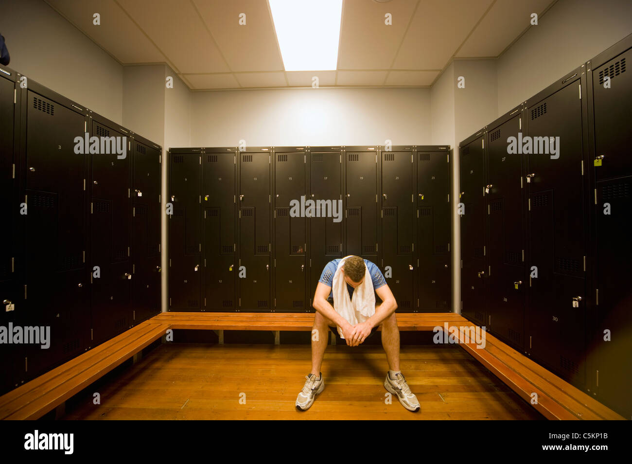 A man sitting in locker room at a gym with a towel round his neck, looking down and looking exhausted Stock Photo