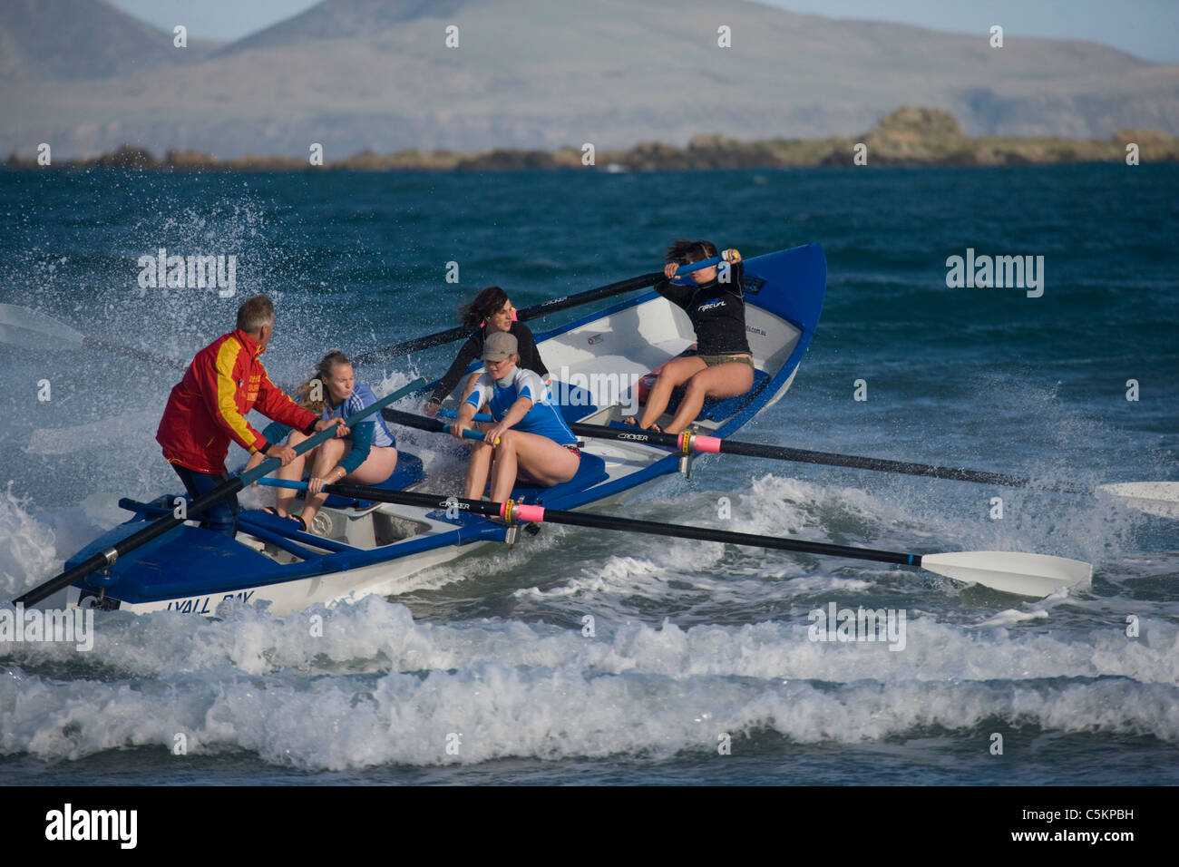 Surf rescue rowing boat going out over breaking waves with four women rowing and a man on rudder, Lyall Bay, Wellington, New Stock Photo