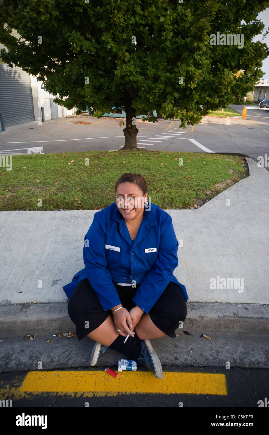 Young Polynesian woman sitting on kerbside outside factory smoking cigarette, wearing blue overall with word 'Temp' embroidered Stock Photo