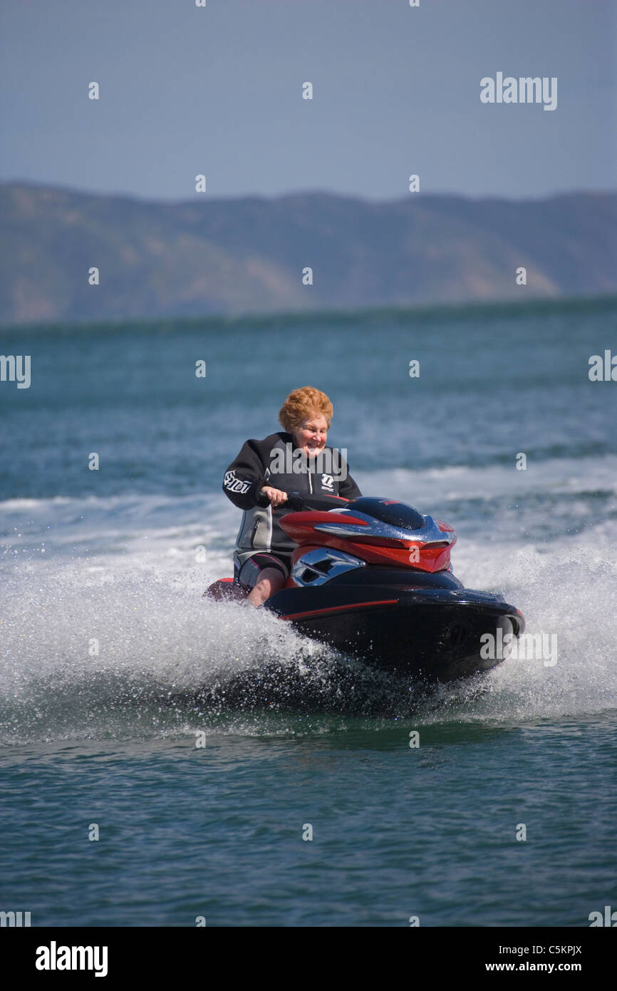 Woman in her 60s driving a jet-ski at speed, wearing a wetsuit, Wellington, New Zealand Stock Photo