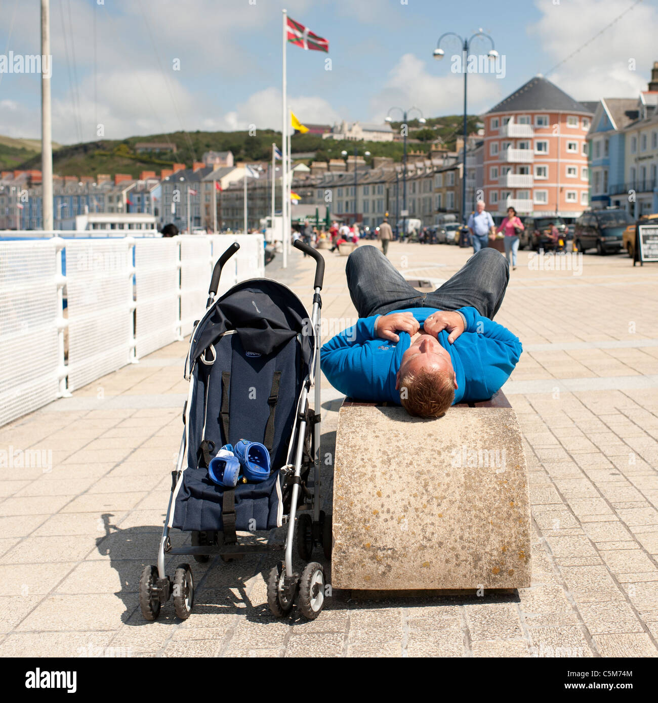 a man lying down on a seaside bench next to an empty kids buggy, aberystwyth wales uk Stock Photo