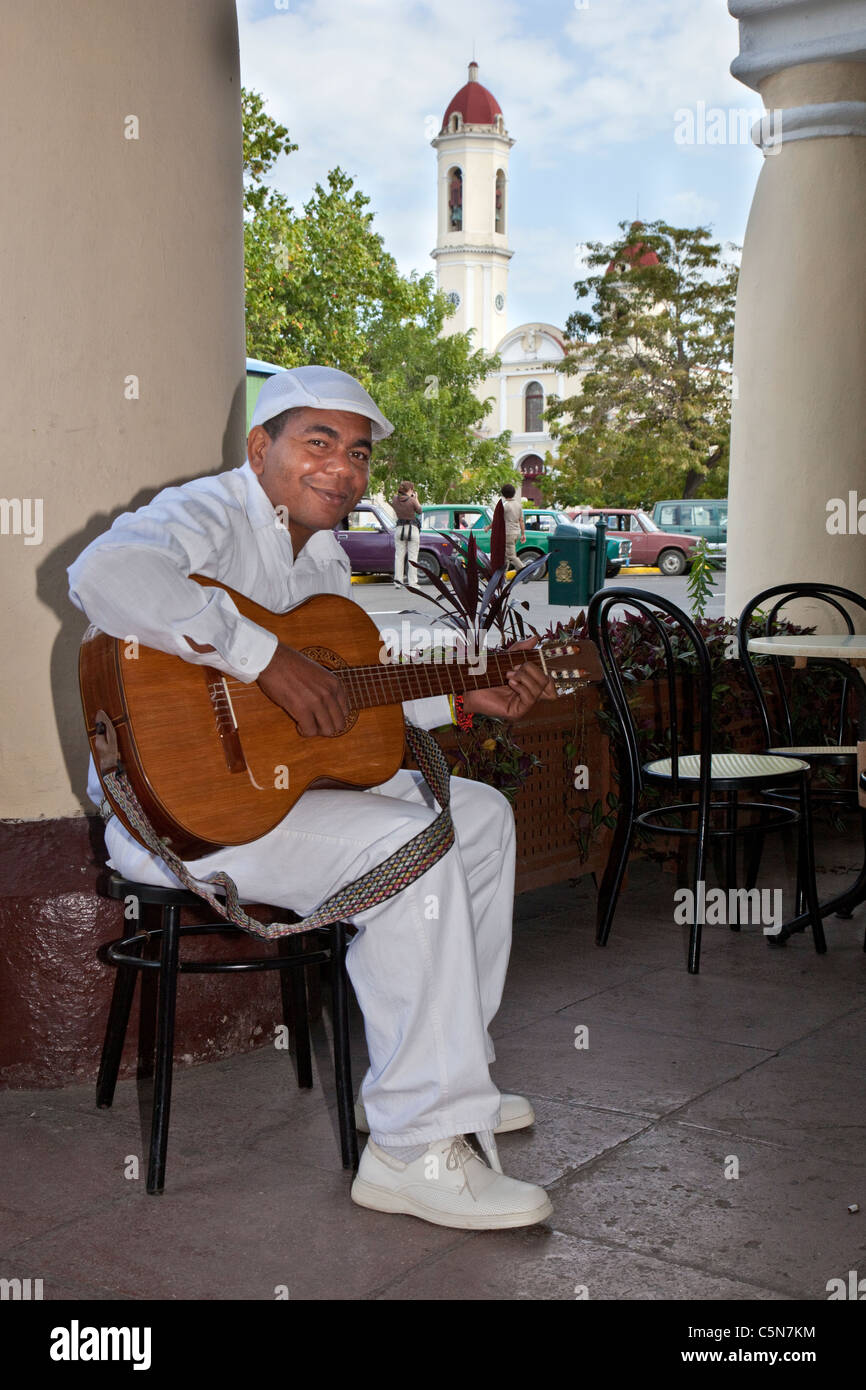 Cuba, Cienfuegos. Afro-Cuban Guitar Player outside a Cafe. Church of La Purisima Concepcion in Background. Stock Photo