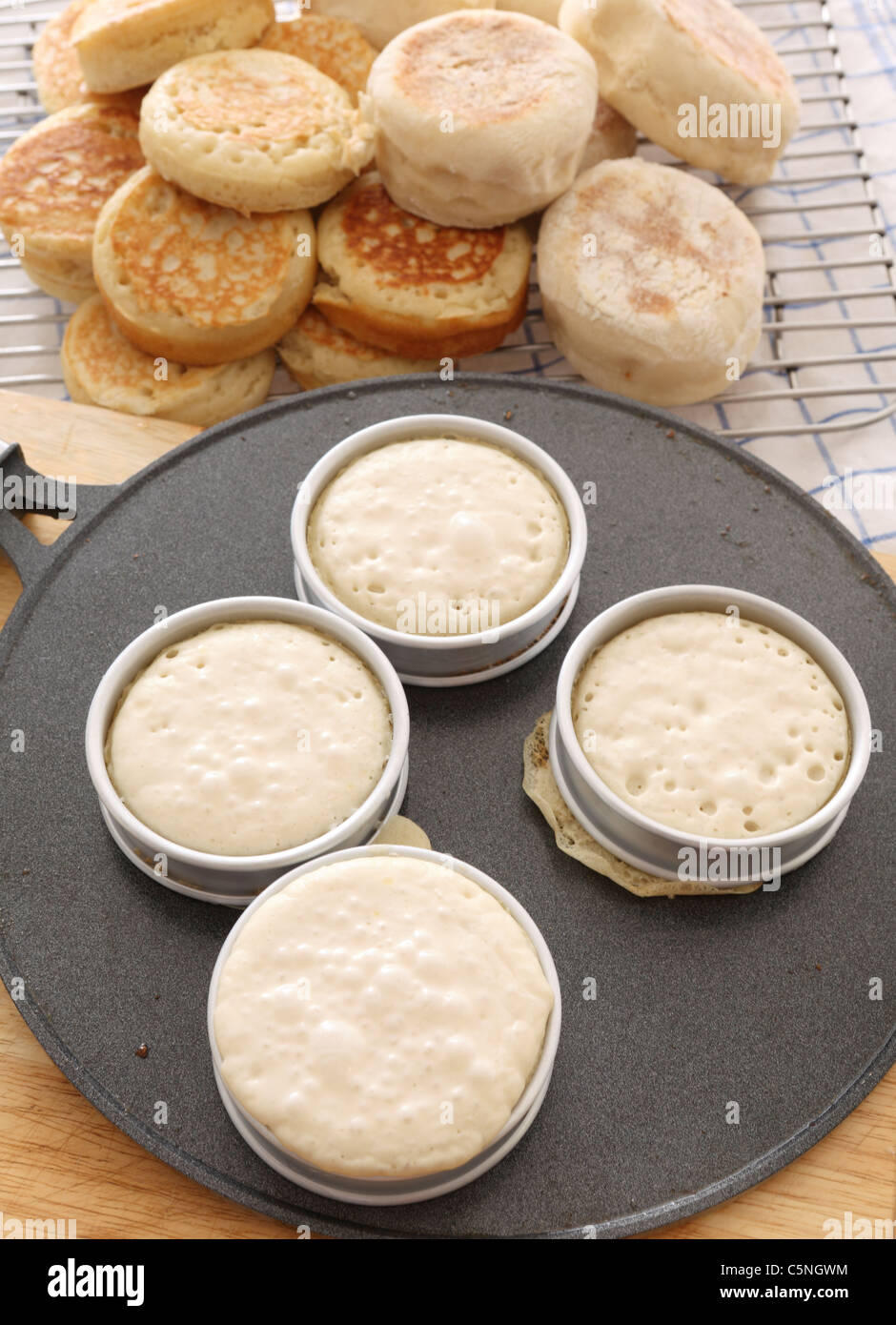 Baking traditional English crumpets in rings on a hot griddle, with a pile of homemade crumpet and muffins in the background Stock Photo