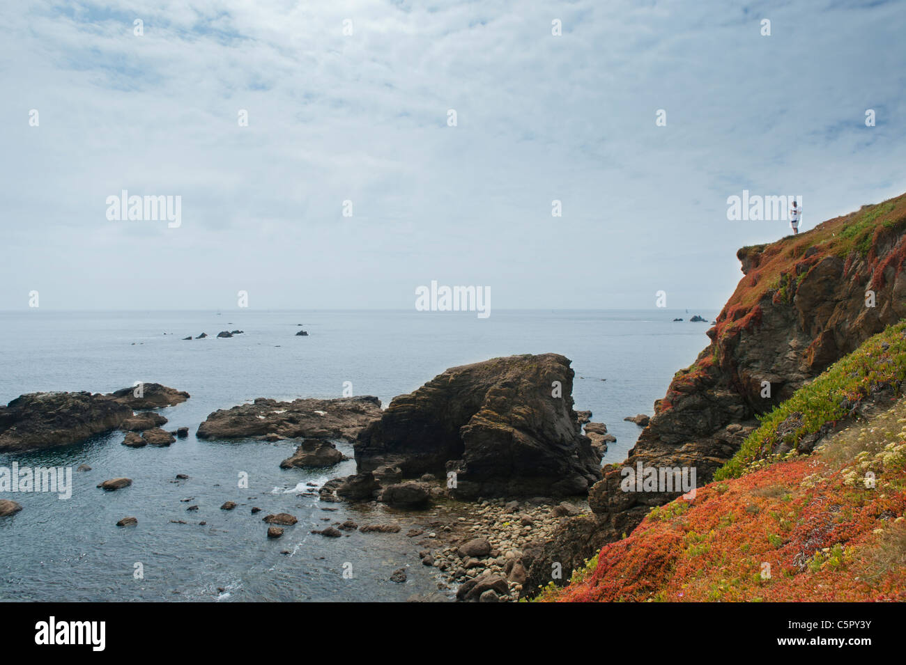 Rocks meet the sea at the coast at Lizard Point, Cornwall, watched by a man stood on a nearby cliff. Stock Photo
