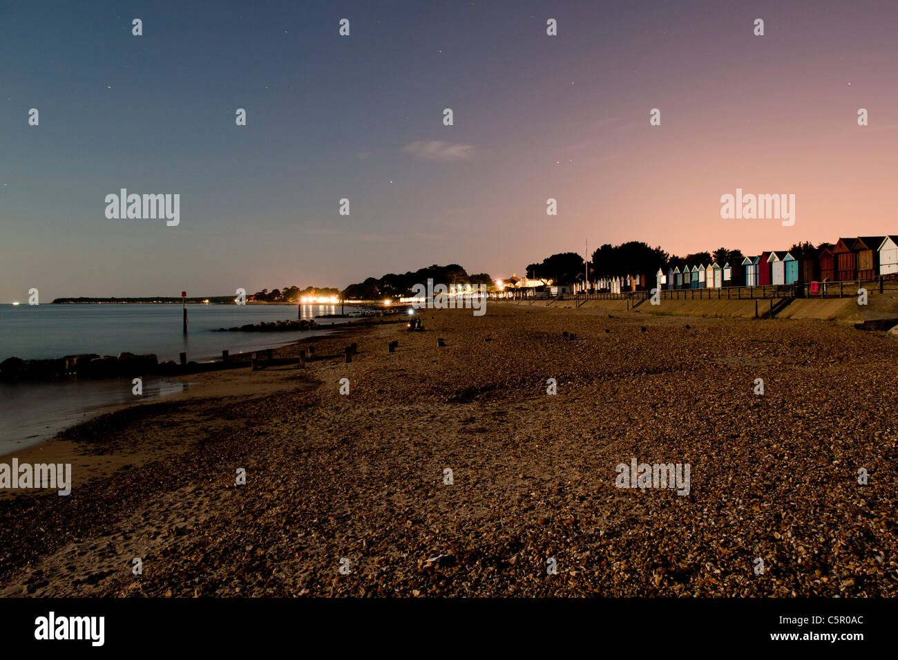 Beach huts are visible against a dimly lit night sky on a pebble beach near to Highcliffe, Dorset. Stock Photo