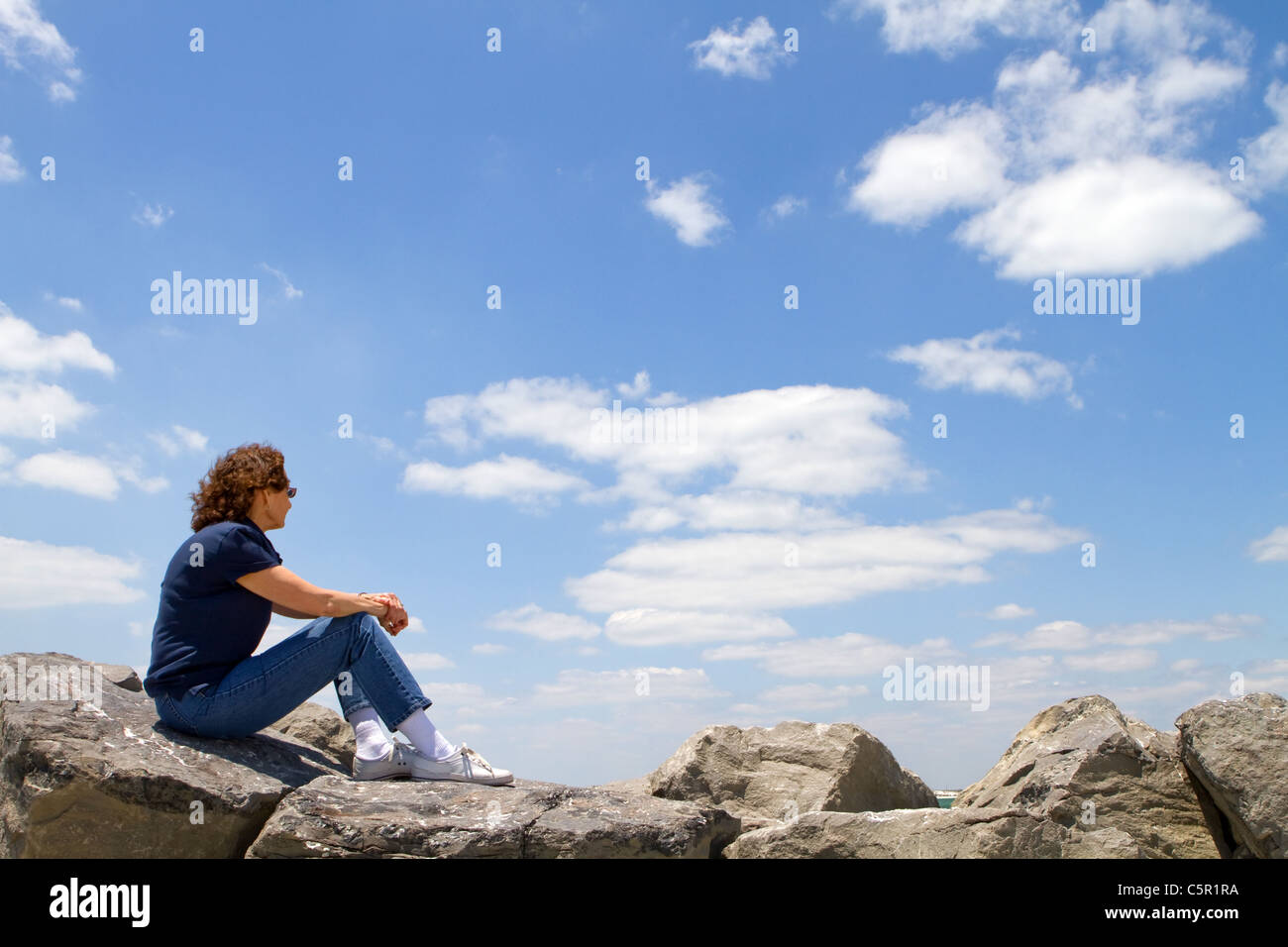 Meditating sixty year old mature woman sits on rocks and gazes out into a cloudy blue sky. Stock Photo