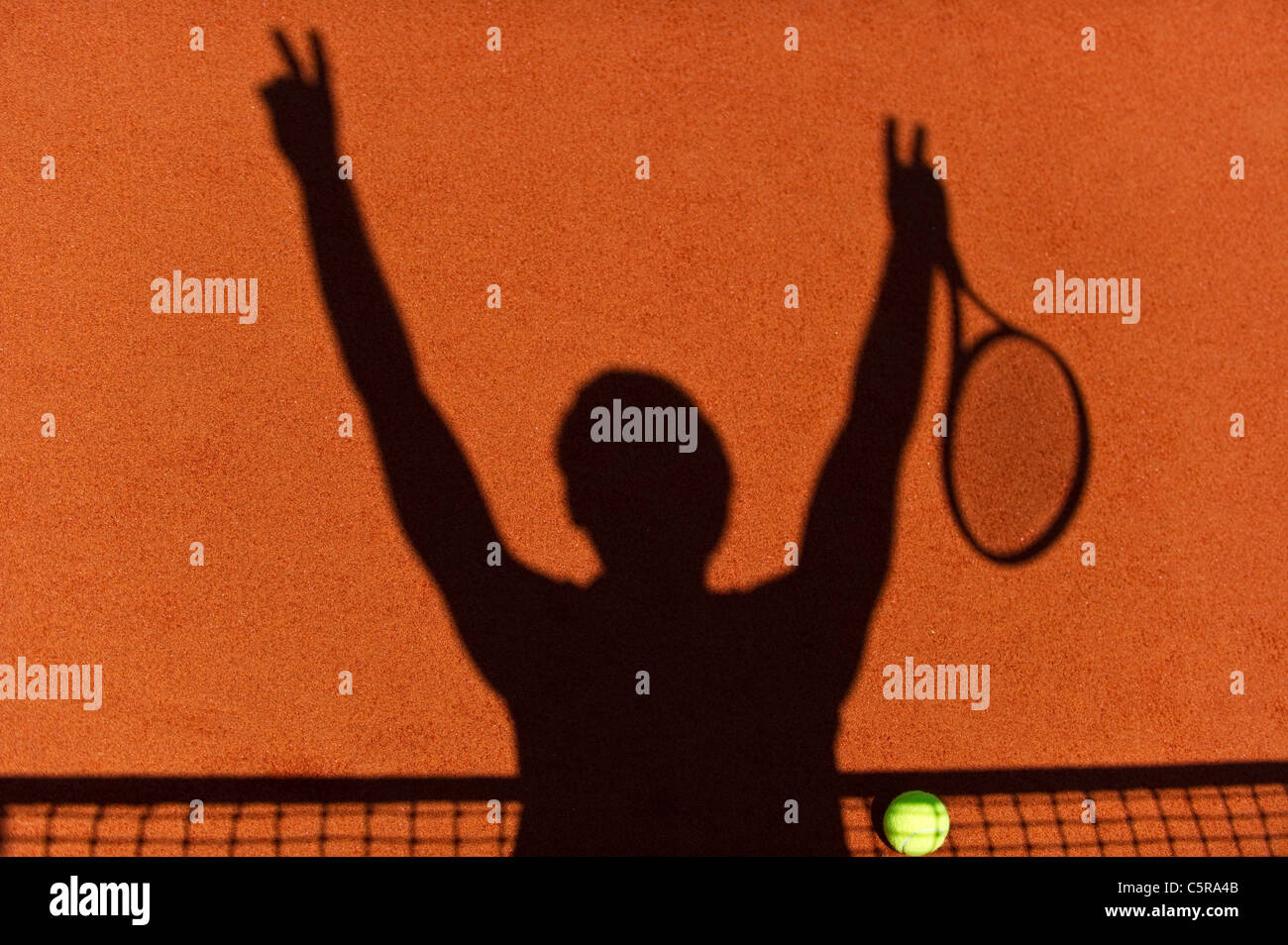 A tennis players silhouette at the net celebrates victory with tennis ball on court. Stock Photo