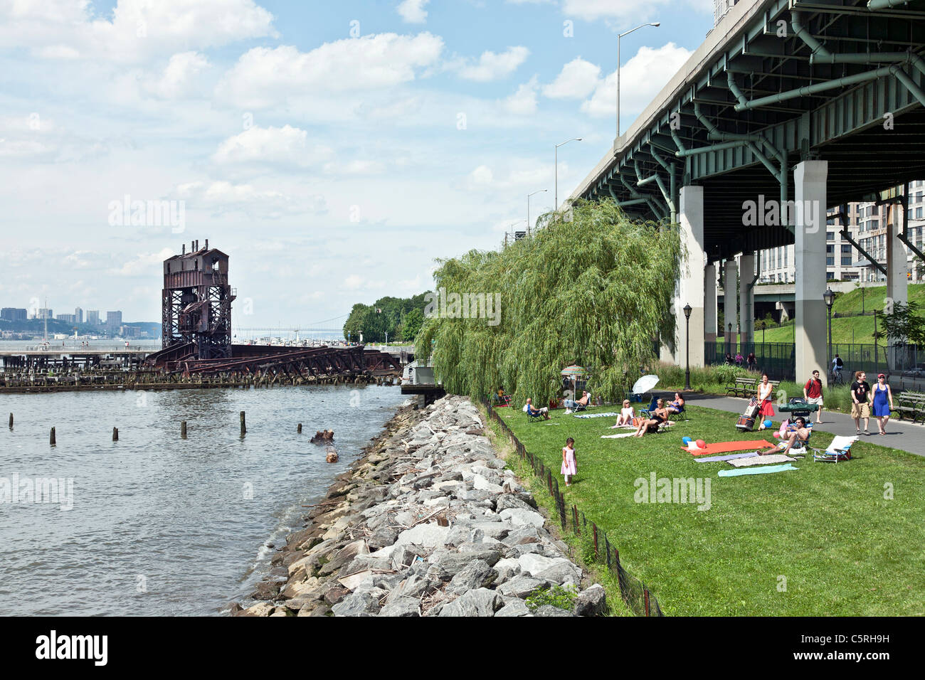 families & friends sunning on lawn at waters edge next to elevated highway while strollers & bicycles pass in Riverside park NY Stock Photo