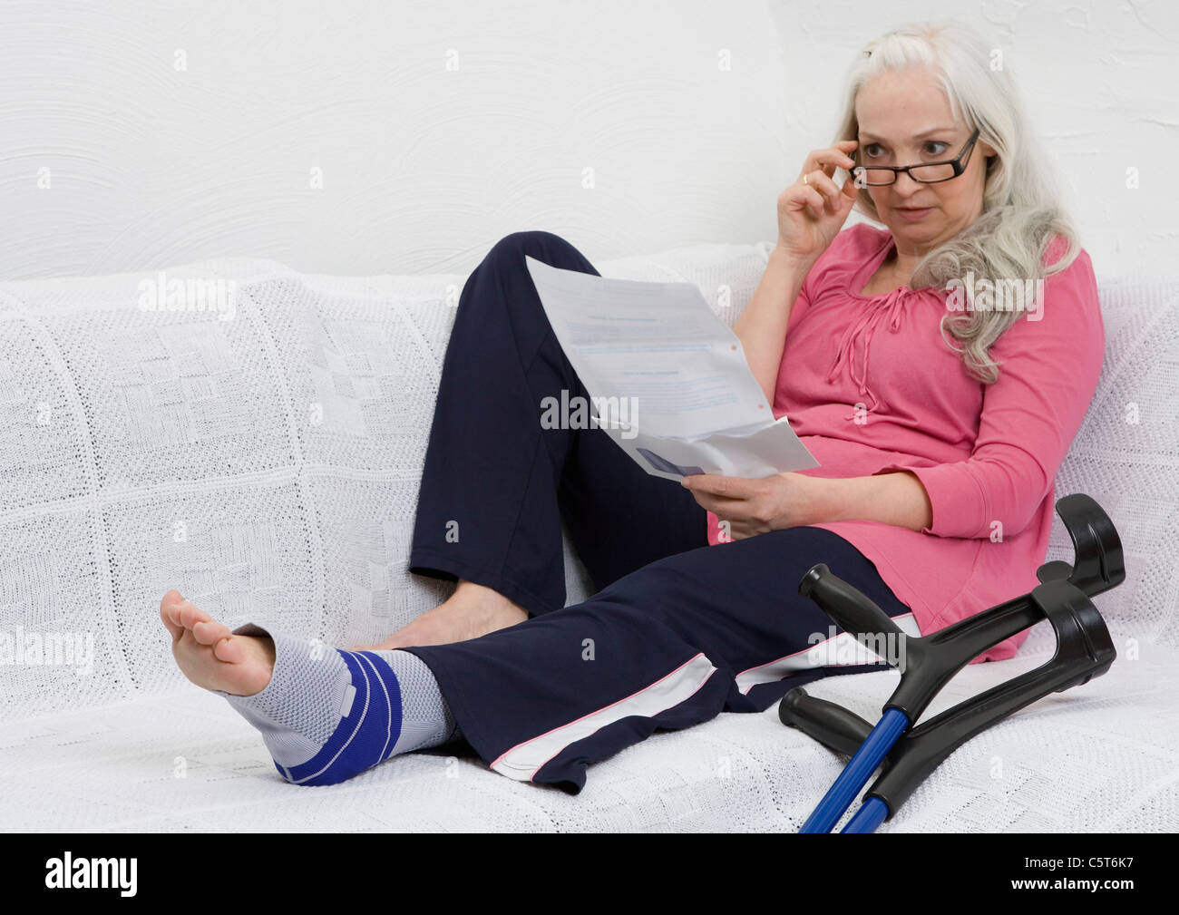 Germany, Duesseldorf, Woman with bandage reading letter on sofa besides crutches Stock Photo