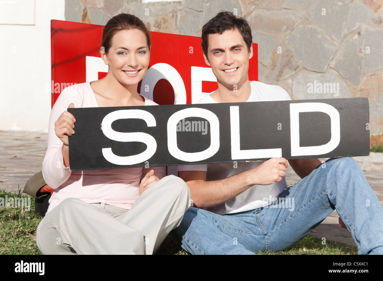Happy couple sitting against their new house with sold sign Stock Photo ...