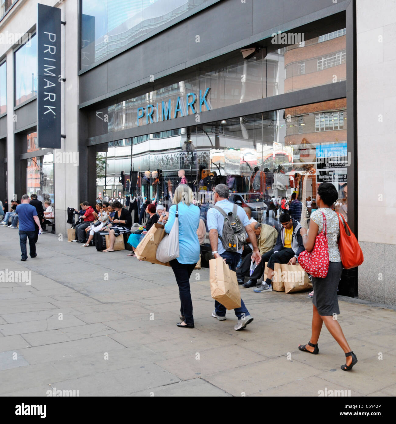 Shoppers with brown paper shopping bags walking past or sitting on window sill Primark clothing shop Oxford Street scene London West End England UK Stock Photo
