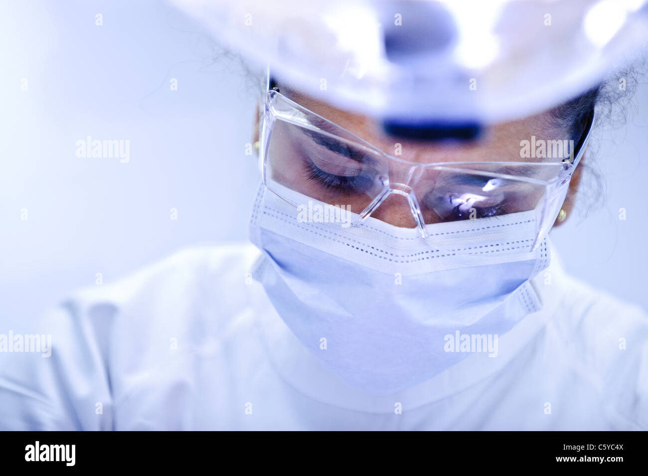 Close up of face of young female dentist wearing a surgical mask and goggles and white dental tunic looking down Stock Photo