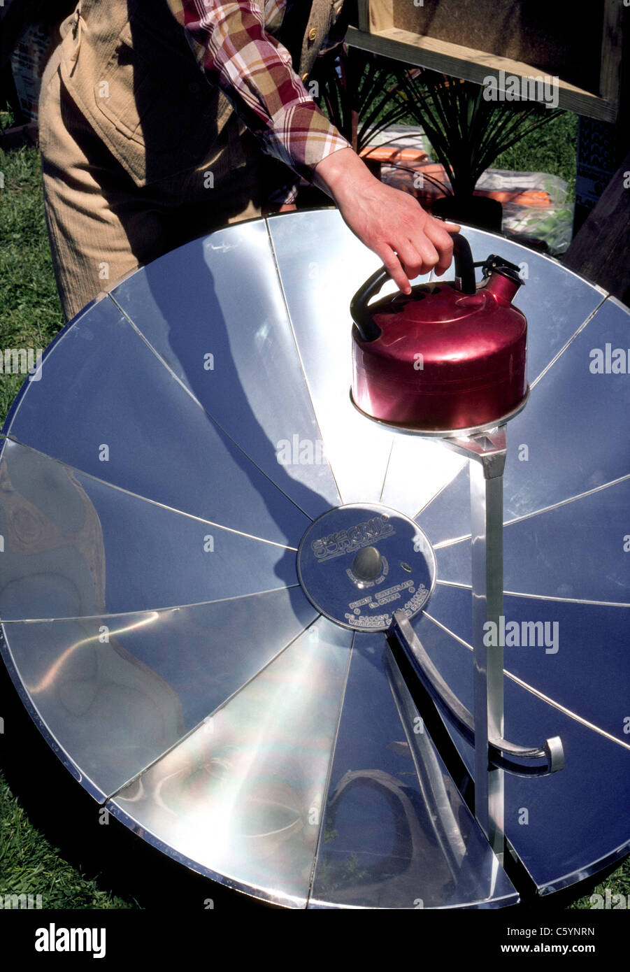 The parabolic reflector of a portable solar cooker focuses energy from the heat of the sun onto the bottom of a teakettle to boil water in New Zealand. Stock Photo