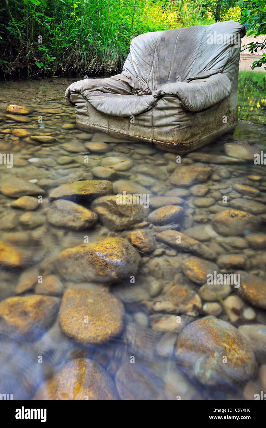 Fly tipping. An old armchair illegally dumped in a clear woodland stream. Stock Photo