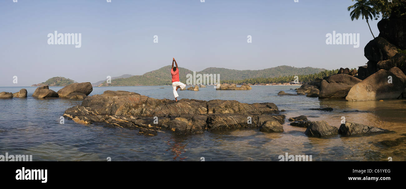 An Indian man does yoga on the rocks of the bay at at the popular tourist resort destination of Palolem Beach, Goa state, India Stock Photo