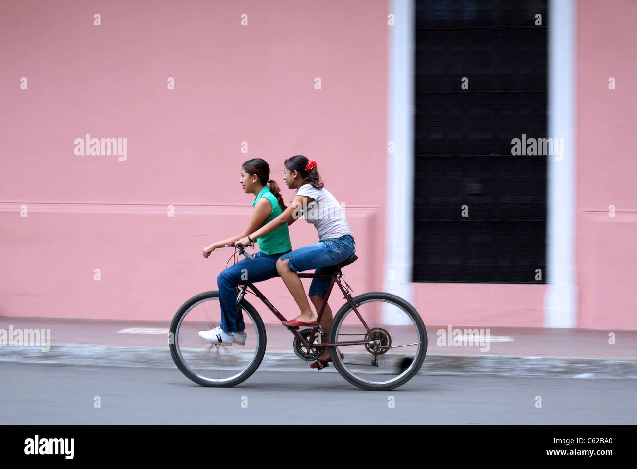 Girls riding bicycle past pink building. Stock Photo