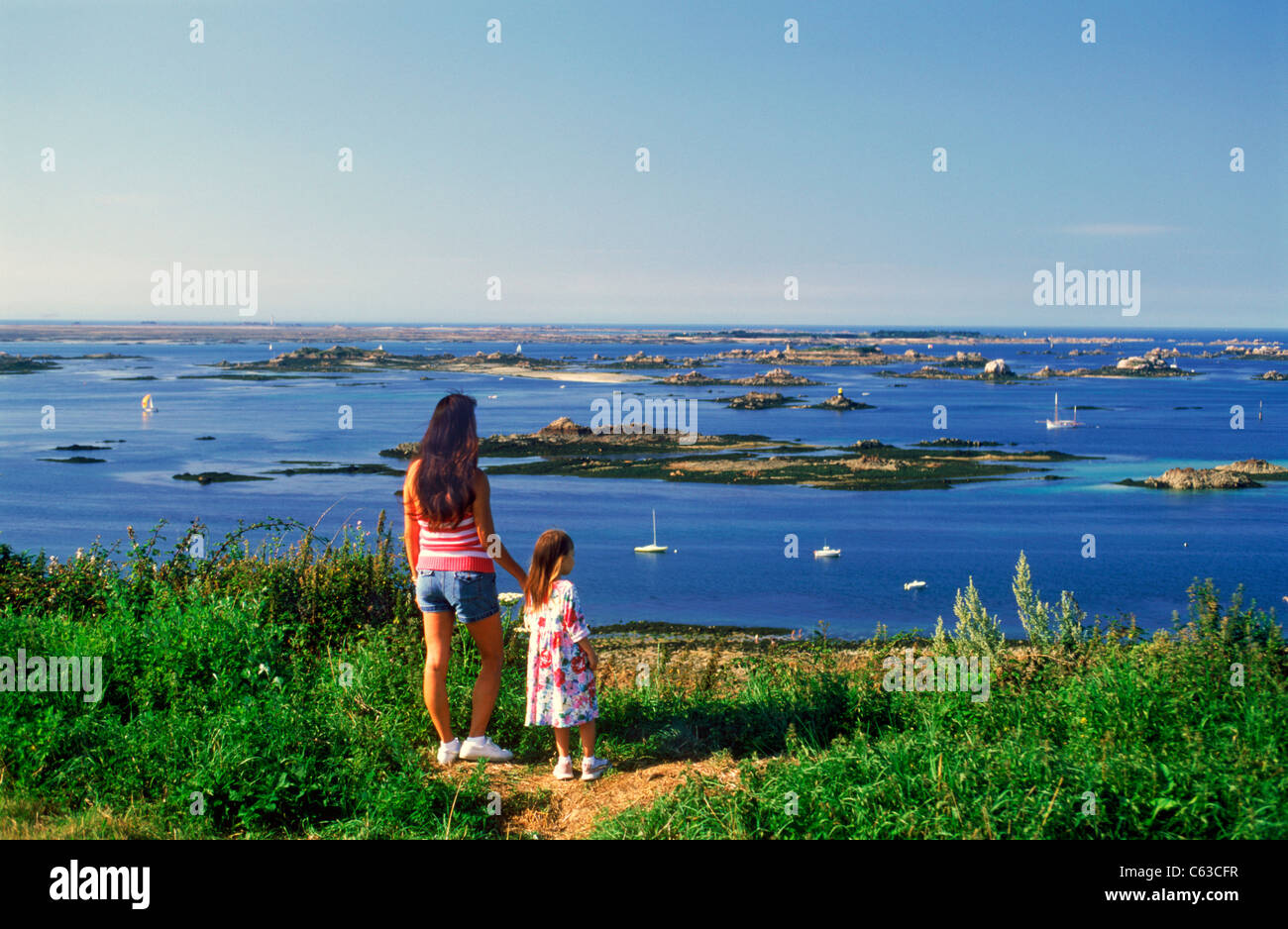 Mother and daughter standing on bluff overlooking small islands and sailboats along Brittany Bretagne Coast in Northern France. Stock Photo