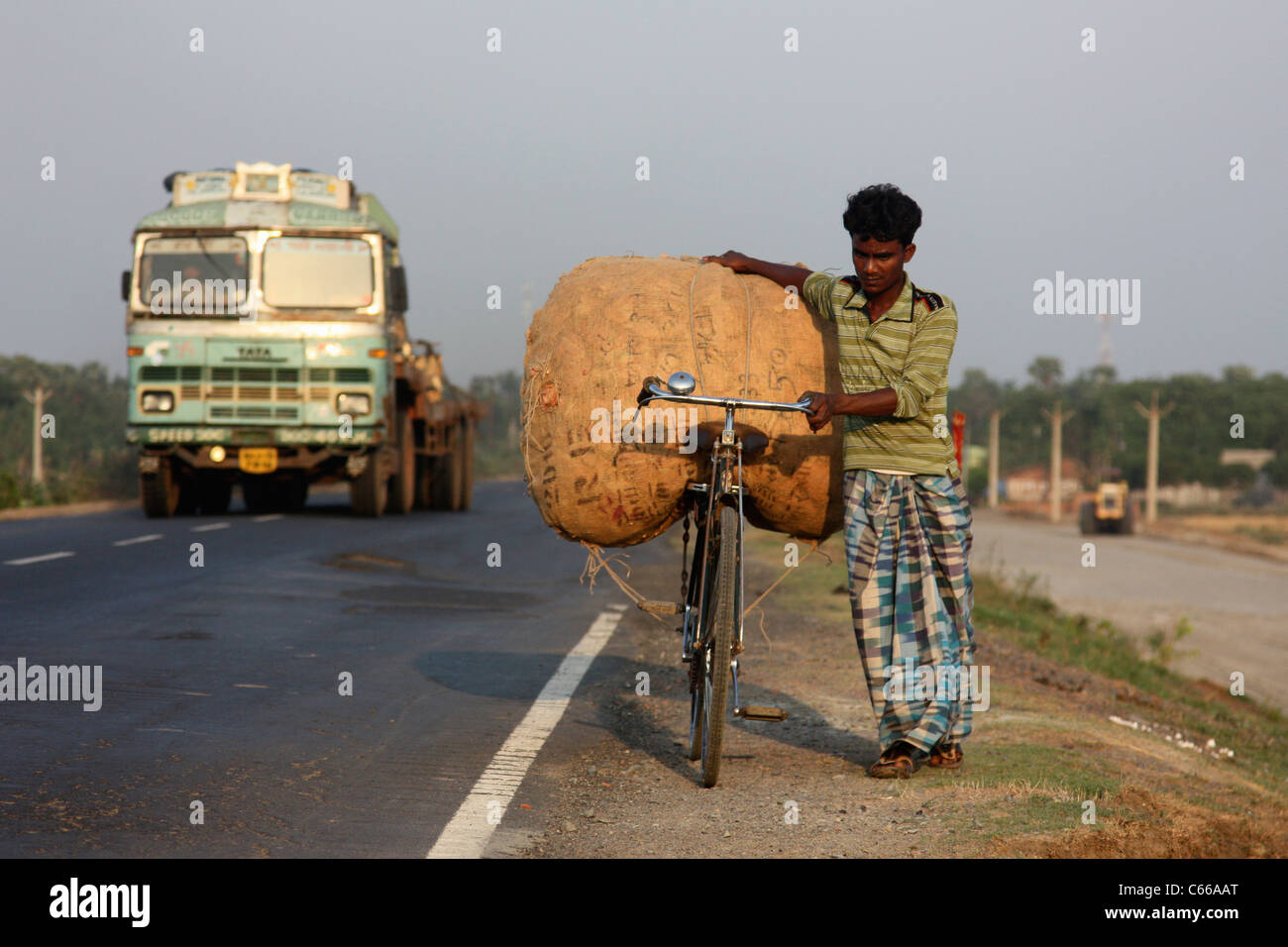 Young man struggles with heavy sack on bicycle up a busy highway at sunset in West Bengal India Stock Photo