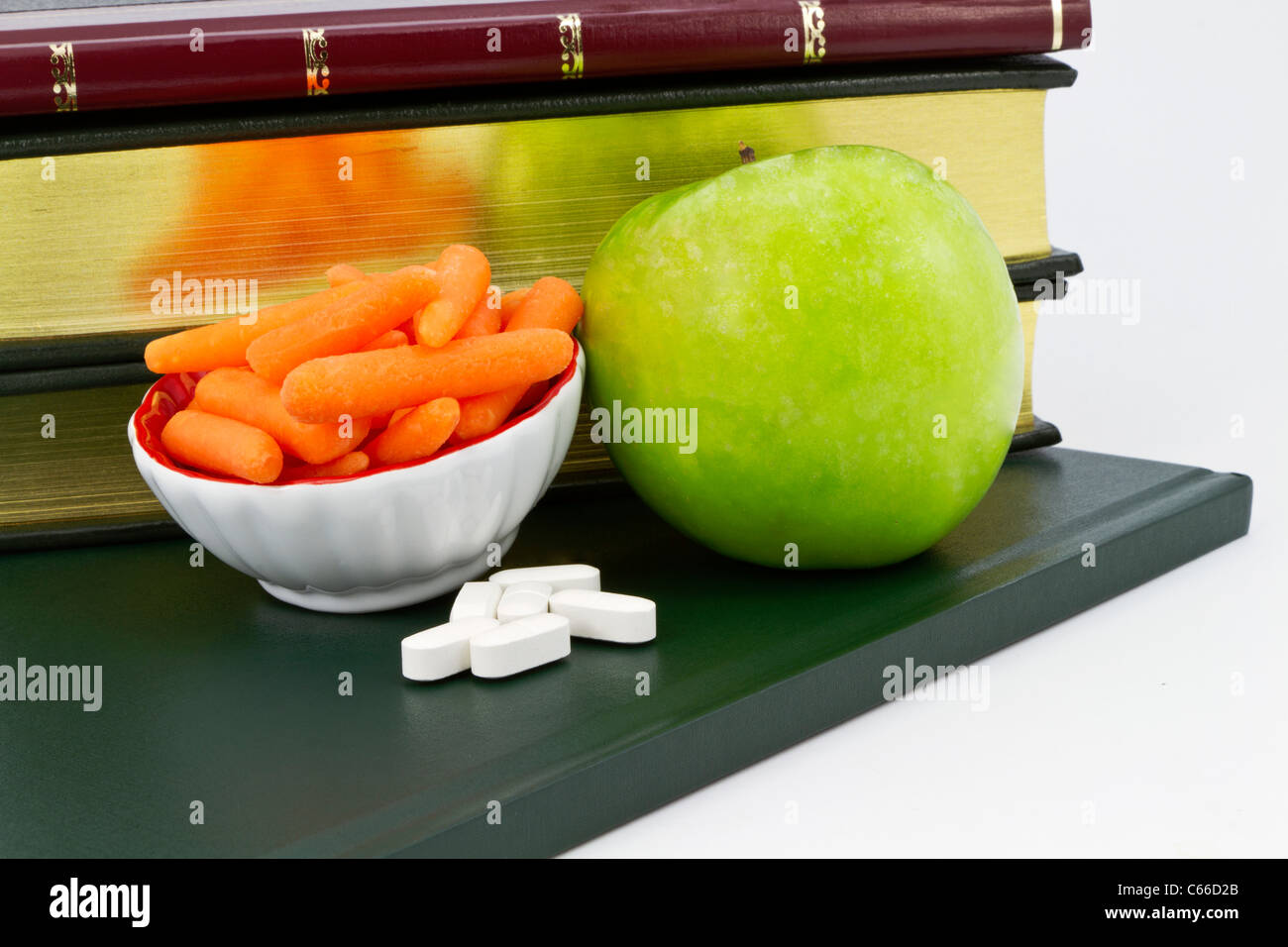 Healthy carrots and a green apple placed with calcium supplement pills with books behind. Stock Photo