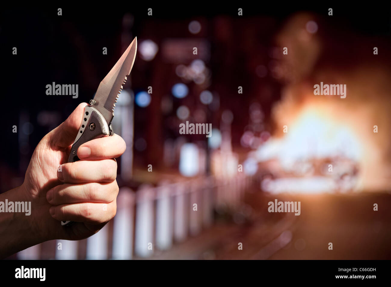 Knife Crime / Riots / Violence Concept. Hand holding a knife with a serrated edge while a car burns in the background. London UK Stock Photo