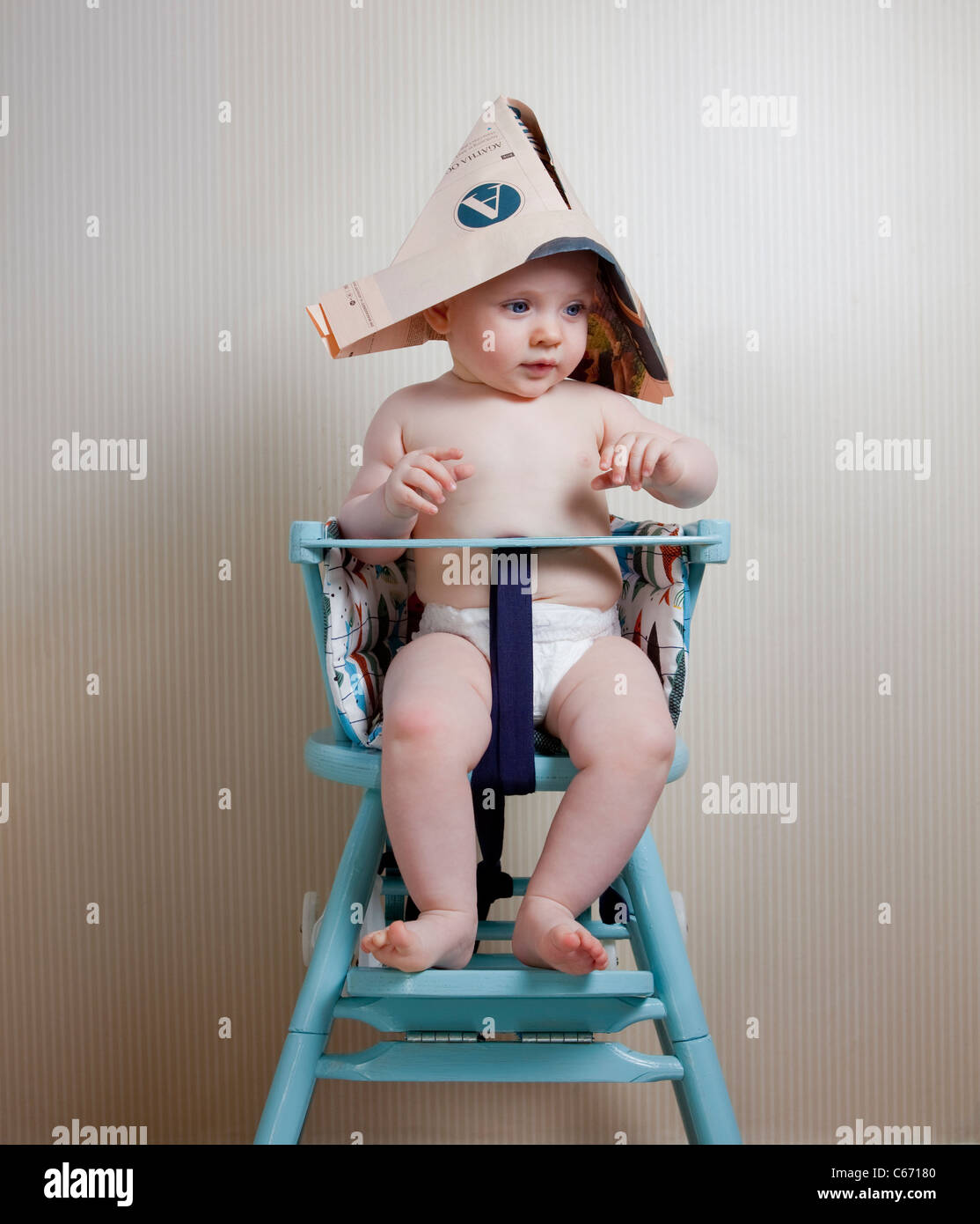 6 months old baby sitting in baby chair wearing a newspaper hat Stock Photo
