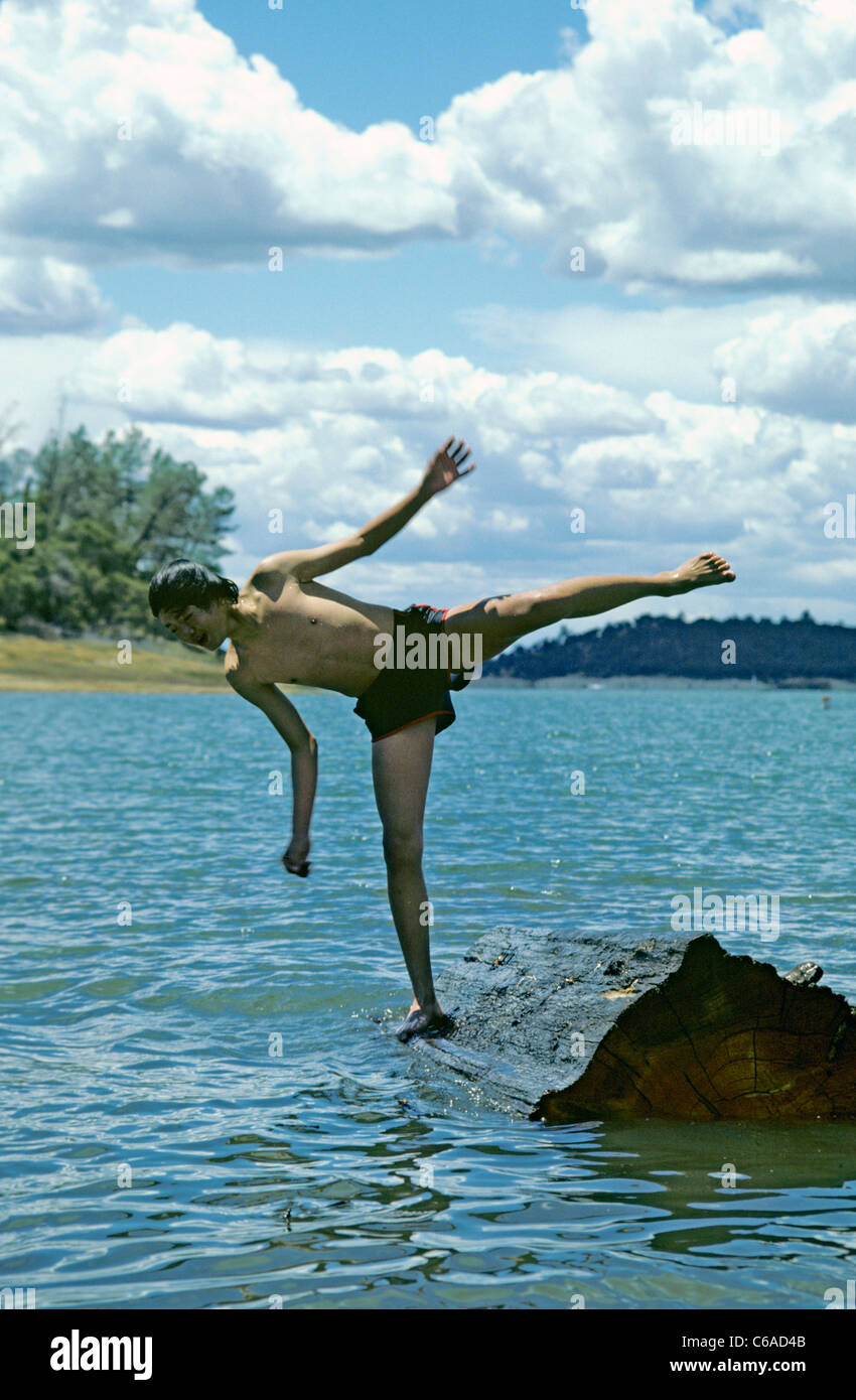 Asian teen balances on floating log in blue lake during long hot summer ...