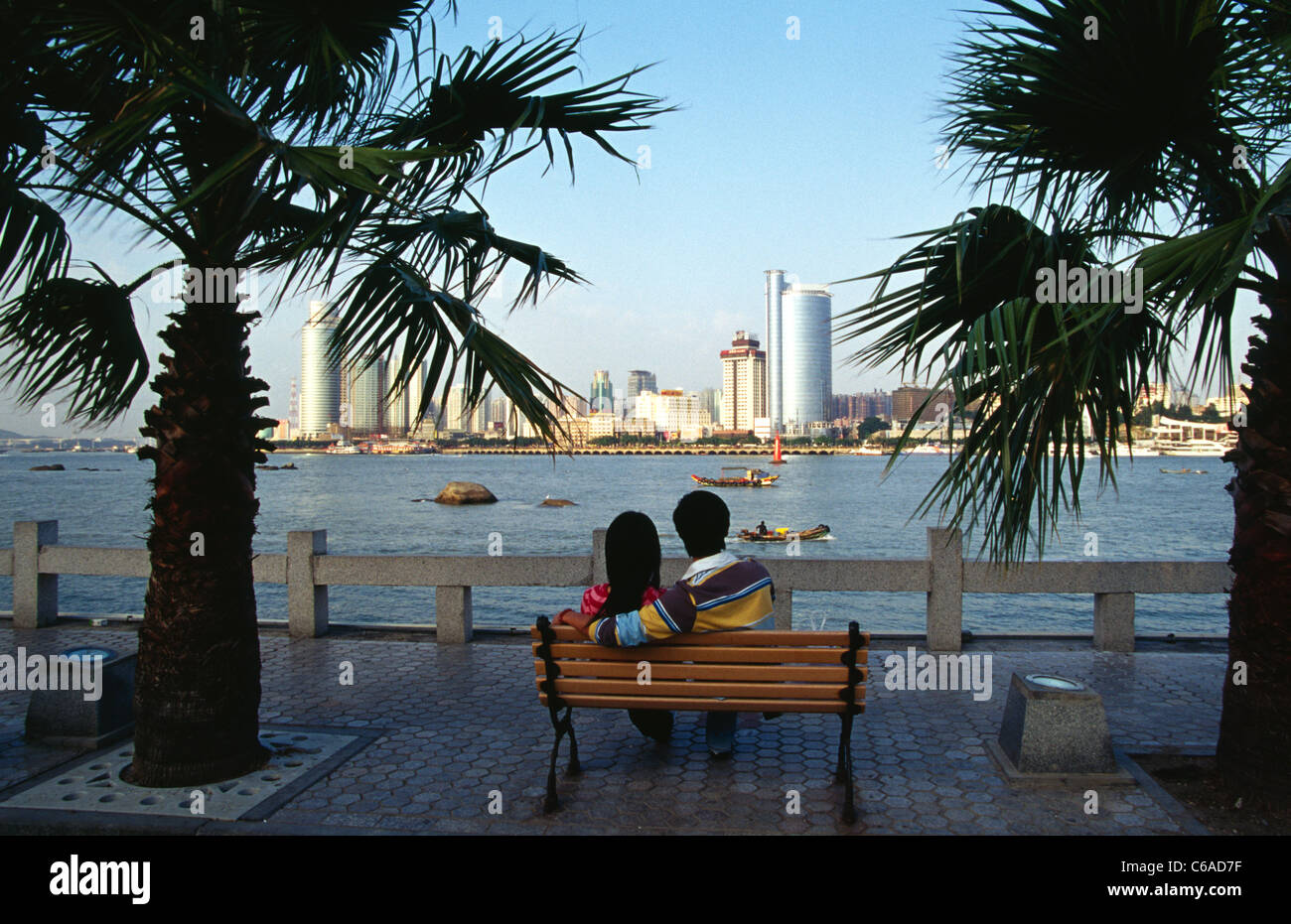 a young couple sit on bench looking at skyline of Xiamen China from tourist island of Gulangyu China Stock Photo