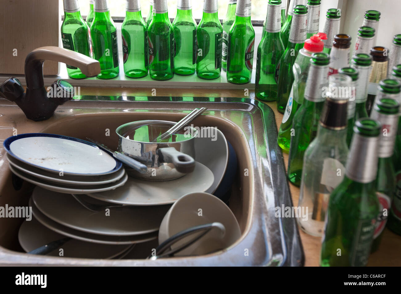 A kitchen sink full of dirty dishes and plates, surrounded by numerous empty green beer bottles. Stock Photo